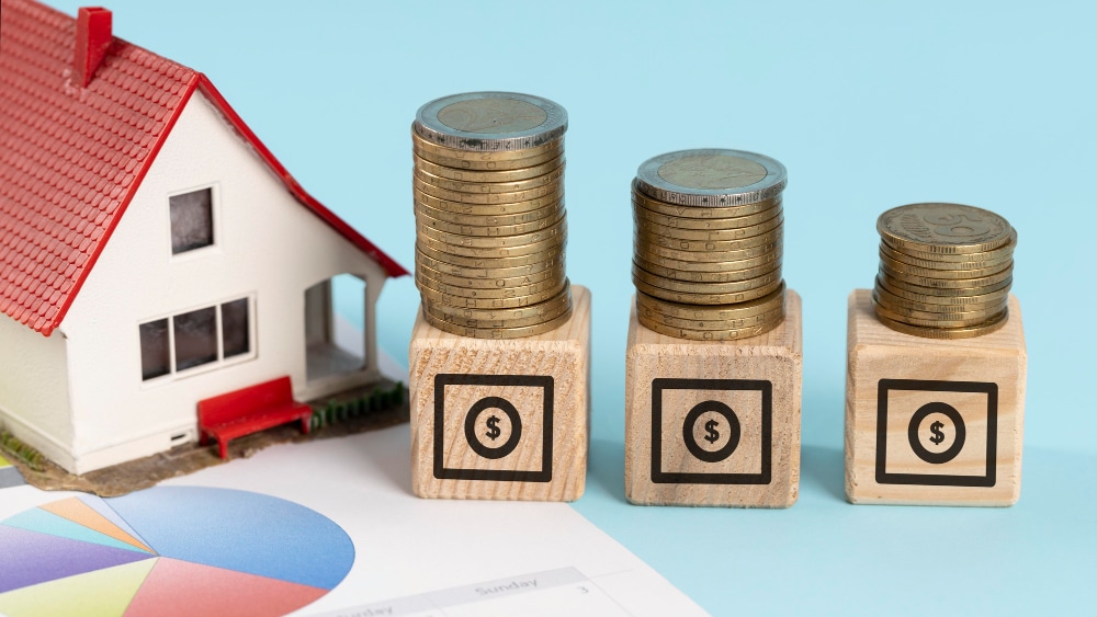 Imagen de una casa con dados de madera que simbolizan dinero y pilas de monedas, representando el riesgo de que te puedan embargar todo el dinero de la cuenta bancaria.