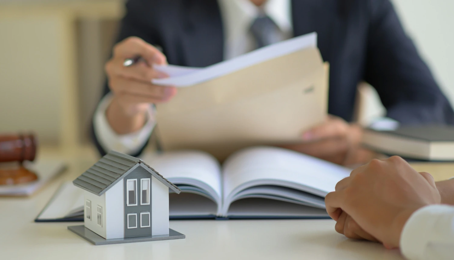 Hombre con tarje y corbata sentado en una mesa con documentos en la mano, un libro sobre la mesa y la maqueta de una casa en referencia a explicar a sus clientes qué es una ejecución hipotecaria y cómo enfrentarse a una oposición a la ejecución hipotecaria para evitar el embargo de su casa