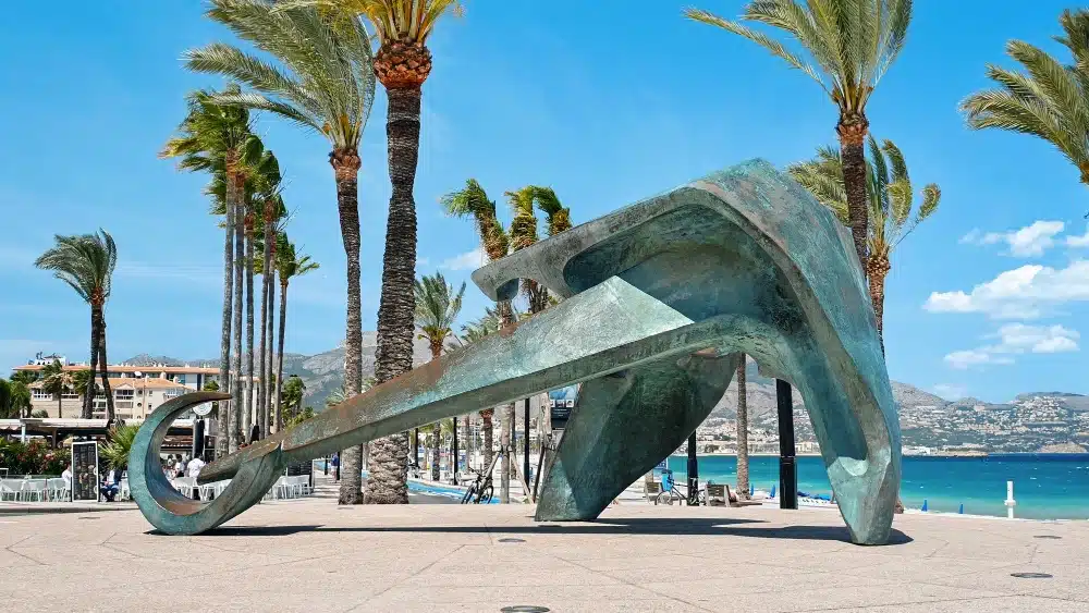 Escultura del ancla en el Paseo Marítimo de Altea, con palmeras y mar de fondo, cerca de las oficinas de Asoban Abogados en Mallorca.