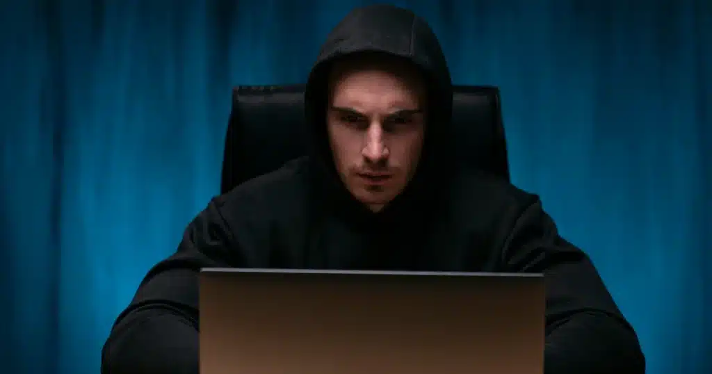 Hombre con sudadera negra y capucha frente a un portátil cometiendo un delito de estafa online.