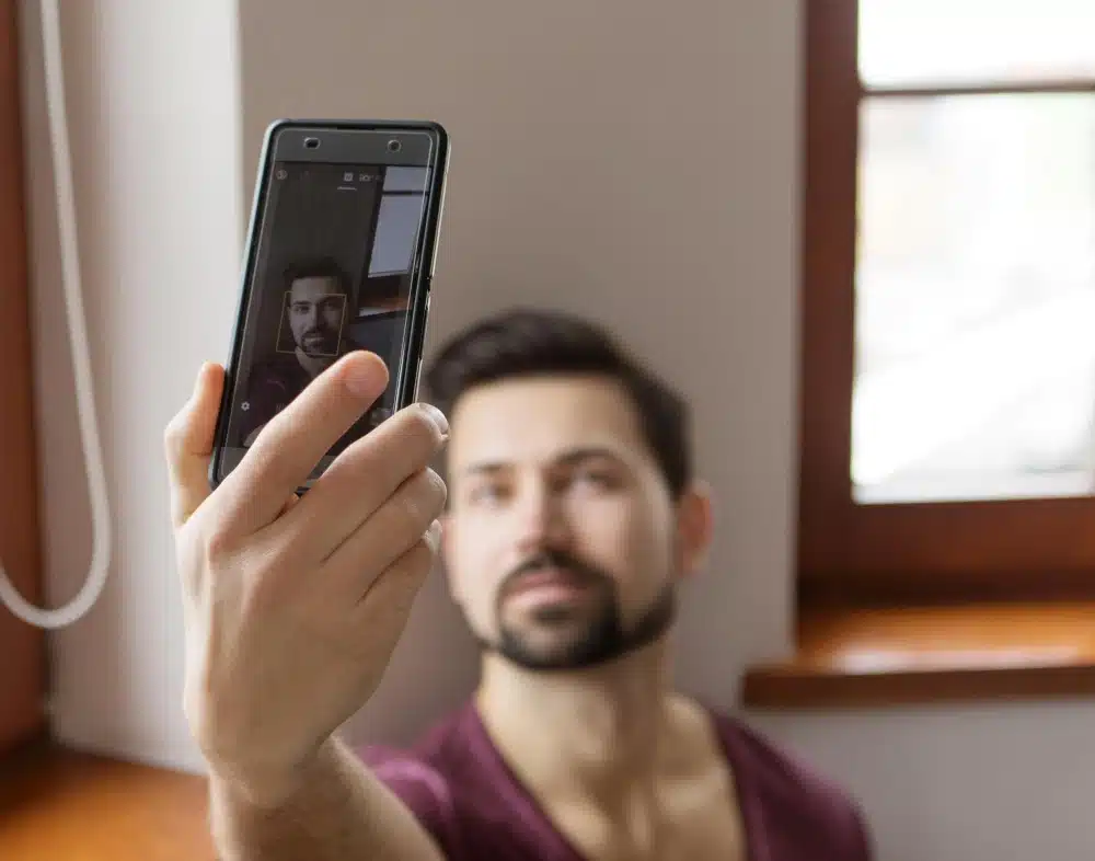 Hombre haciéndose una selfie con el móvil, simulando la creación de fotos típicas usadas por timadores del amor en perfiles falsos.
