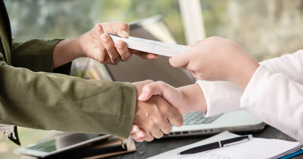 Dos personas estrechando la mano mientras se entrega dinero en efectivo, simbolizando un acuerdo financiero transparente. Fidinda es legal y segura.