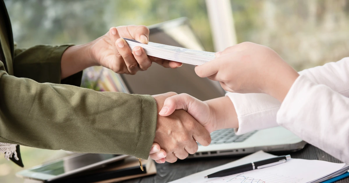 Dos personas estrechando la mano mientras se entrega dinero en efectivo, simbolizando un acuerdo financiero transparente. Fidinda es legal y segura.