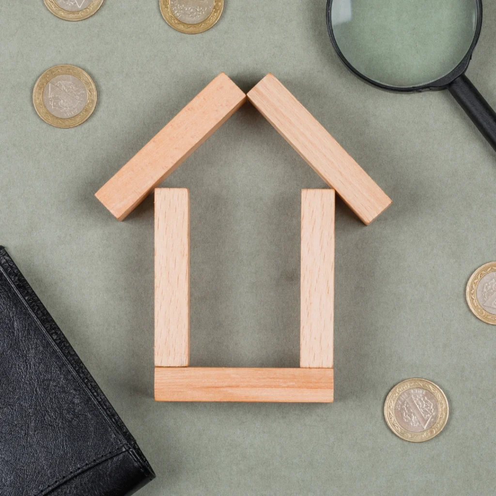 Casa hecha con bloques de madera, rodeada de monedas de euro, una lupa y una cartera negra, representando el análisis del Banco de España sobre el IRPH en hipotecas, tipos de interés y condiciones financieras de préstamos hipotecarios.