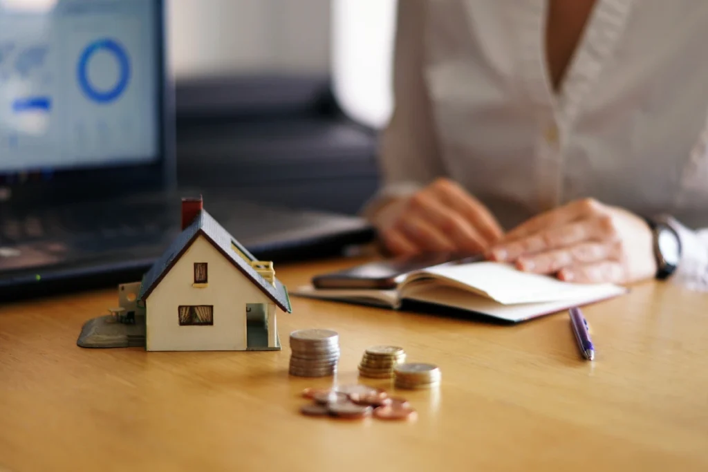 Persona revisando documentos financieros junto a una casa en miniatura, monedas y un portátil, representando el análisis de condiciones ofrecidas por bancos con hipotecas IRPH en el mercado español.