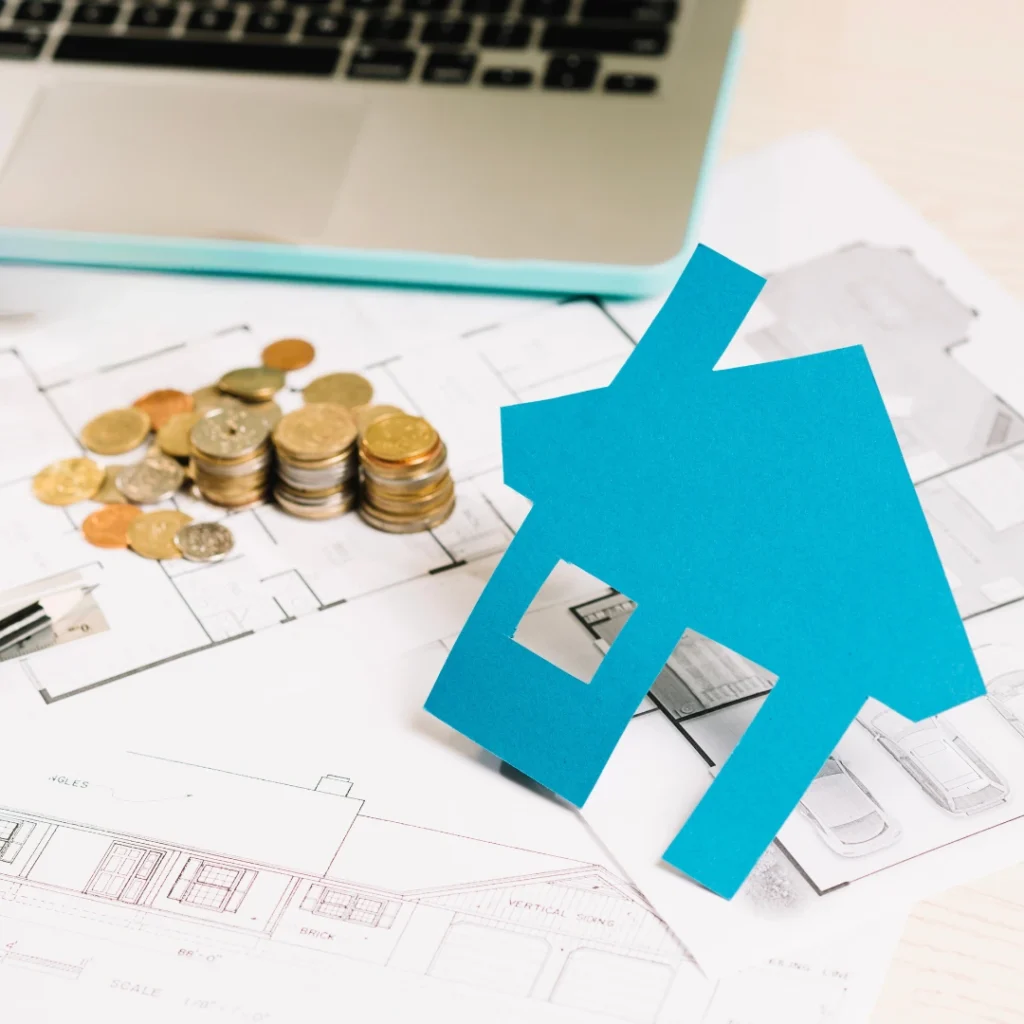 Monedas apiladas, planos arquitectónicos y un símbolo de casa junto a un ordenador, representando el Índice de Referencia de Préstamos Hipotecarios (IRPH).