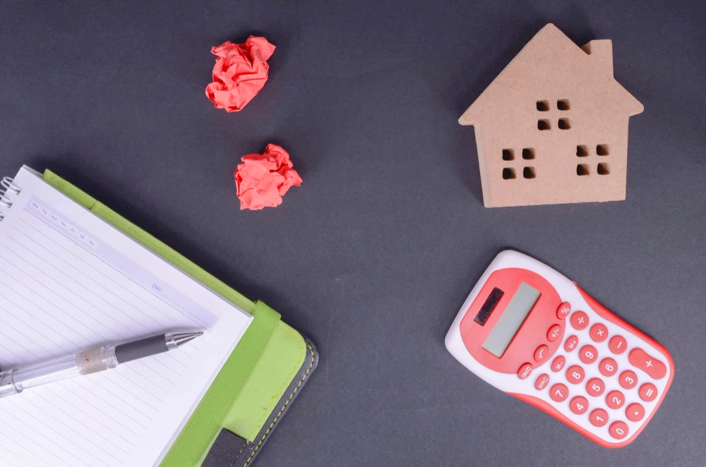 Calculadora, libreta y figura de una casa sobre fondo gris que representan el cálculo de qué gastos de hipoteca se pueden reclamar al banco.
