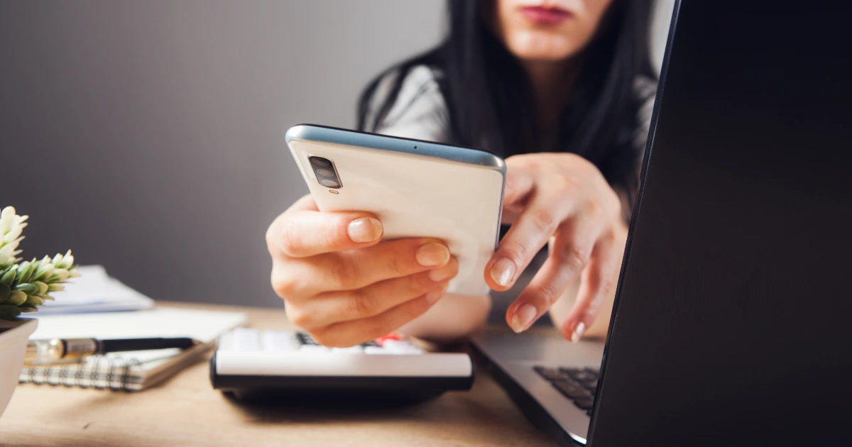 Mujer utilizando su teléfono móvil y ordenador, representando un caso de smishing o fraude por SMS, una ciberestafa común que busca robar datos personales mediante mensajes fraudulentos.