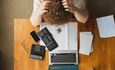 Persona preocupada revisando facturas y documentos en la mesa, intentando evitar ser incluido en un fichero de morosos, calculadora y ordenador portátil en el escritorio.